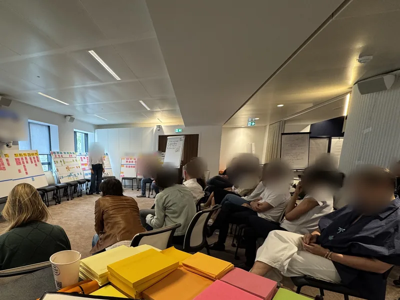 Atelier de discovery : des participants assis face aux murs couverts de parcours utilisateurs et de post-its. Les visages et les textes sont floutés.